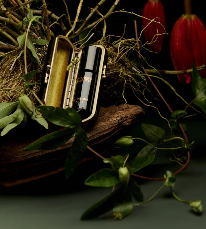 Alt: Vintner’s Daughter serum bottle in a luxurious velvet case, placed among natural vines, wood, and red flowers on a dark green background.